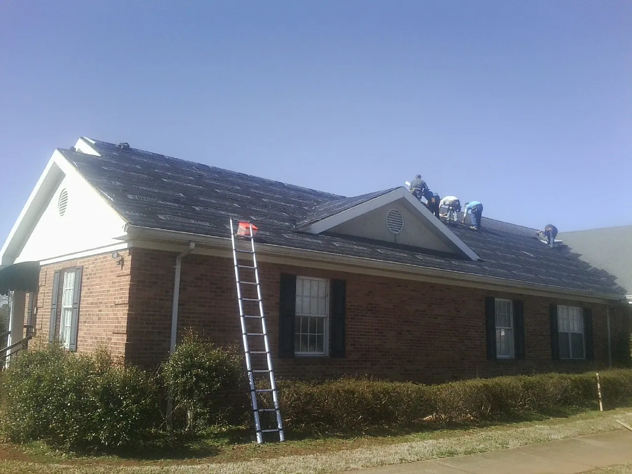Crew tearing shingles off a brick ranch roof with a ladder set — Metal Roof Repair work by our crew in Maynard